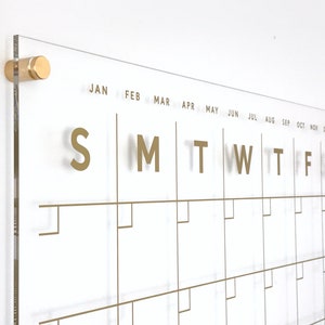 Op de afbeelding: Een transparante acrylkalender met gouden letters en een gouden frame. De kalender heeft de dagen van de week met "S M T W T F S" en de maanden van het jaar met "JAN FEB MAR APR MEI JUN JUL AUG SEP OKT NOV DEC" gelabeld.