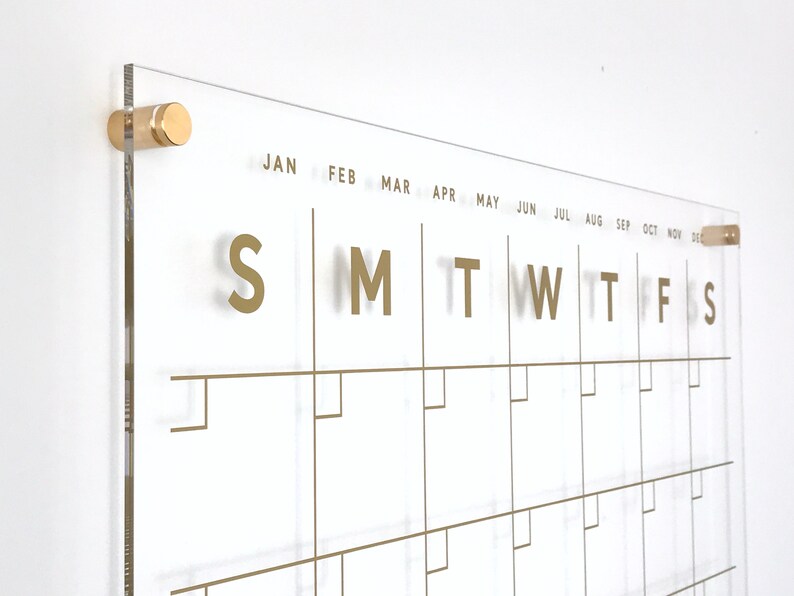 Op de afbeelding: Een transparante acrylkalender met gouden letters en een gouden frame. De kalender heeft de dagen van de week met "S M T W T F S" en de maanden van het jaar met "JAN FEB MAR APR MEI JUN JUL AUG SEP OKT NOV DEC" gelabeld.