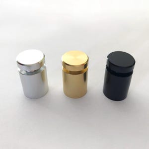 Puede incluir: Tres soportes de exhibición cilíndricos de acrílico en plata, oro y negro. Cada soporte tiene un borde elevado alrededor de la parte superior e inferior.