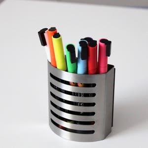 Op de afbeelding: Een zilveren metalen pennenhouder met een gebogen ontwerp bevat een verscheidenheid aan kleurrijke stiften. De stiften hebben zwarte dopjes en zijn verticaal gerangschikt.