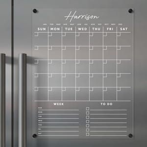 Puede incluir: Un calendario de borrado en seco de acrílico transparente con un marco blanco y texto blanco. El calendario tiene una vista mensual con espacios para cada día de la semana. También tiene una vista semanal con espacios para cada día de la semana y una lista de tareas pendientes. El calendario está etiquetado como "Harrison".