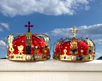 May include: Two ornate gold crowns with red velvet interiors, adorned with faux jewels and pearls. The crowns feature intricate filigree designs and are topped with crosses. The background is a bright blue sky with fluffy white clouds.