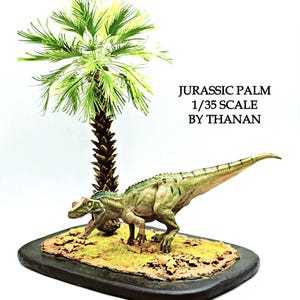Peut inclure: Une maquette à l'échelle 1/35 d'un dinosaure théropode vert et beige sur une base brune texturée à côté d'un palmier. Le texte JURASSIC PALM 1/35 SCALE BY THANAN est imprimé en noir sur le côté droit de la présentation du modèle.