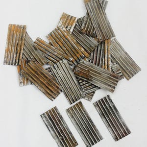 Op de afbeelding: Een verzameling kleine, rechthoekige metalen stukken met een gegolfde textuur. De stukken hebben een verweerde uitstraling met tinten zilver, bruin en roest, wat wijst op een verouderde of industriële esthetiek. De opstelling is verspreid over een wit oppervlak.