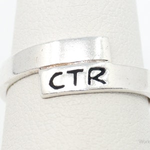 May include: Silver ring with a band that wraps around the finger twice. The top of the ring has a rectangular piece with the letters "CTR" engraved in black.