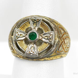 May include: A gold and silver-toned ring featuring a Celtic cross design with a green gemstone center. The ring has intricate knotwork detailing on the band and cross, showcasing a vintage aesthetic. The ring is displayed on a white surface.