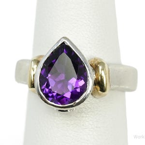 May include: A silver and gold ring featuring a pear-shaped, deep purple amethyst gemstone. The ring has a silver band with gold accents on either side of the gemstone. A classic jewelry piece.