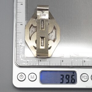 May include: A silver-toned metal money clip with an ornate design, marked "NICKEL SILVER." The clip is on a digital scale, displaying a weight of 39.6 grams. The clip has a geometric shape with decorative cutouts.