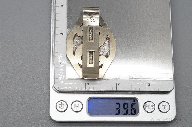 May include: A silver-toned metal money clip with an ornate design, marked "NICKEL SILVER." The clip is on a digital scale, displaying a weight of 39.6 grams. The clip has a geometric shape with decorative cutouts.
