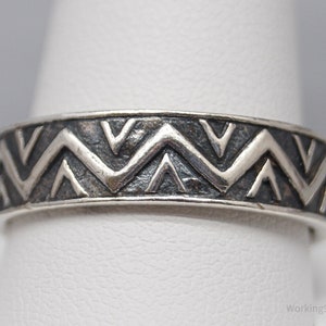 May include: A silver band ring with a geometric pattern of repeating triangles. The ring is on a white display stand.