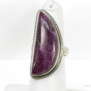 May include: A large, silver-toned ring featuring a striking, elongated, half-moon-shaped purple stone. The stone is framed by a textured, rope-like border. The ring is displayed on a white surface, showcasing its elegant design.