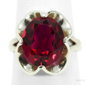 Puede incluir: Un anillo de plata con una gran gema roja oscura de corte ovalado. La gema está engastada en un engaste de plata con forma de flor. El anillo se muestra sobre un fondo blanco, resaltando el diseño y el color de la joya.