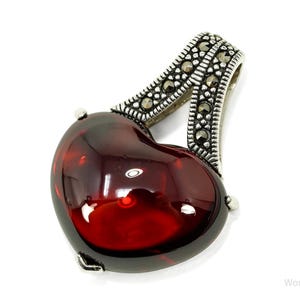 May include: A heart-shaped pendant with a deep red, glossy stone. The pendant has a silver-coloured bail, decorated with small, dark stones. The jewellery piece is set against a white background.