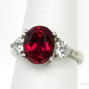 May include: A silver ring featuring a large, oval-cut, deep red gemstone flanked by two smaller, pear-shaped, clear gemstones. The ring is set on a white background, showcasing the jewelry's design and sparkle.