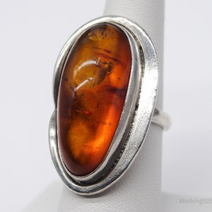 Könnte beinhalten: Ein Silberring mit einem großen, ovalen Bernstein. Der Bernstein hat eine warme, durchscheinende orangebraune Farbe mit sichtbaren Einschlüssen. Der Ring hat ein schlichtes Design mit einem Silberband und einer Lünettenfassung, die die natürliche Schönheit des Bernsteins hervorhebt.