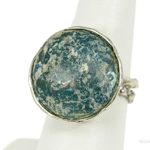 Puede incluir: Anillo de plata con una piedra grande, redonda y moteada en tonos turquesa, azul y gris. La piedra está engastada en un bisel de plata texturizada. El anillo se muestra sobre una superficie blanca.