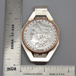 May include: A silver money clip with a Morgan silver dollar coin from 1896. The coin is set in a copper and silver frame. The clip is approximately 2 inches long. The background is a gray surface.