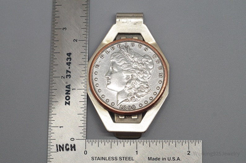 May include: A silver money clip with a Morgan silver dollar coin from 1896. The coin is set in a copper and silver frame. The clip is approximately 2 inches long. The background is a gray surface.