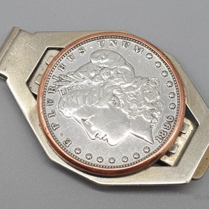 May include: A silver-toned money clip holding a silver coin with a copper-colored rim. The coin features a profile of a woman and the year 1896. The clip has a geometric design.