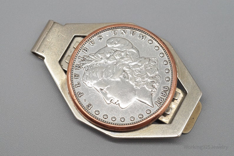 May include: A silver-toned money clip holding a silver coin with a copper-colored rim. The coin features a profile of a woman and the year 1896. The clip has a geometric design.