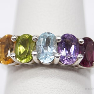 May include: A silver ring featuring five oval gemstones in a row. The gemstones are in shades of yellow, green, light blue, purple, and red. The ring is set against a white background.