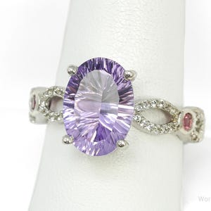 Könnte beinhalten: Ein silberner Ring mit einem großen, oval geschliffenen, hellvioletten Edelstein. Das Band hat ein Unendlichkeitsdesign mit kleinen, klaren Steinen und rosa Edelsteinen. Der Ring wird auf einer weißen Oberfläche präsentiert.