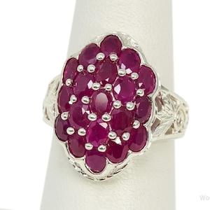 May include: A silver ring featuring a cluster of oval-cut deep pinkish-red stones arranged in a vertical, multi-layered oval formation. The silver band displays intricate, engraved floral-like patterns along the sides of the setting.