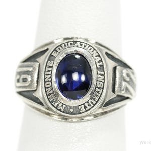 May include: A silver-toned ring with a dark blue oval stone at the center. The ring is engraved with the words "Mennonite Educational Institute" and the numbers "19" and "52". The ring is on a white background.