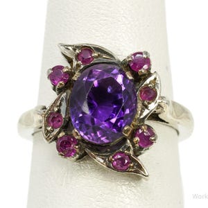 May include: A silver ring featuring a large oval-cut purple amethyst centre stone surrounded by smaller round-cut pink rubies set in a floral-inspired metal arrangement. The text Working925Jewelry is visible in the bottom right corner.