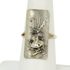 May include: A silver ring featuring a rectangular face with a textured, matte finish. The center of the face displays a raised, polished silver sculptural element resembling a small vessel or pot surrounded by abstract, organic, wire-like forms.