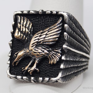 May include: A silver-toned ring featuring a detailed eagle design. The eagle, with gold-colored accents, is set against a textured black background. The ring has a rectangular face with feather detailing on the sides.