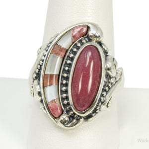 May include: A sterling silver ring featuring a large, oval-shaped, polished pink stone set in a beaded frame. Beside the central stone is a curved inlay section composed of alternating segments of white mother-of-pearl and pink stone.