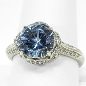 May include: A silver ring featuring a large, light blue gemstone. The gemstone is round and faceted, with a brilliant cut. The ring's band is also silver and is adorned with small, clear accent stones. The ring is set against a white background.