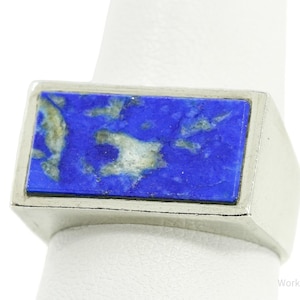 May include: A silver ring featuring a rectangular lapis lazuli stone. The stone is a vibrant blue with streaks of gold and white. The ring has a simple, modern design, showcasing the natural beauty of the gemstone.