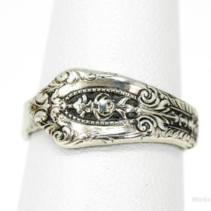 May include: Silver-toned ring with ornate floral and scrollwork details. The ring features a central design with a rose and other floral elements. The ring is displayed on a white surface.
