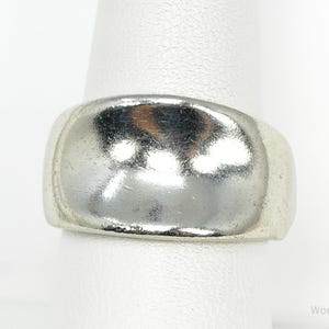 May include: A silver ring with a rounded rectangular face. The ring is displayed on a white surface, highlighting its polished, reflective surface. The ring's design is simple and classic, suitable for everyday wear.