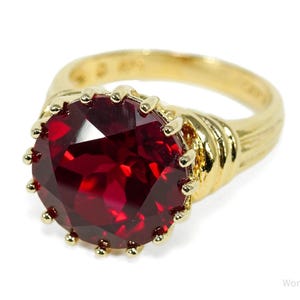 May include: A gold-toned ring featuring a large, faceted, deep red gemstone. The ring has a detailed, textured band and a bezel setting with small gold accents around the gemstone. The ring is set against a white background.