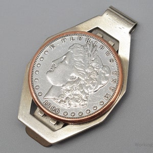 May include: A silver money clip holding a silver coin with a copper-colored rim. The coin features a profile of a woman and the year 1896. The money clip has a geometric design and is set against a gray background.