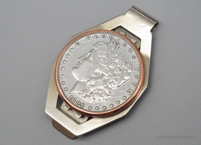 May include: A silver money clip holding a silver coin with a copper-colored rim. The coin features a profile of a woman and the year 1896. The money clip has a geometric design and is set against a gray background.