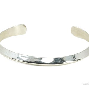 May include: A polished silver cuff bracelet with a smooth, reflective surface and a tapered, open-ended design. The metal band features a subtle, rounded ridge running along the centre, creating a clean, metallic finish with curved ends.