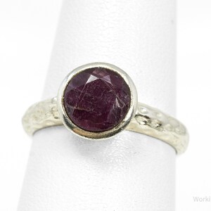 Könnte beinhalten: Ein Silberring mit einem runden, facettierten, dunkelroten Edelstein in einer glatten Fassung. Die Ringschiene hat eine texturierte, gehämmerte Oberfläche. Der Text Working925Jewelry ist in der unteren rechten Ecke des Bildes zu sehen.