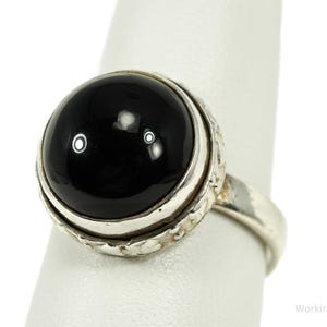 Könnte beinhalten: Silberring mit einem großen, runden, polierten schwarzen Stein. Der Stein ist in einer strukturierten Lünette gefasst, und das Band ist schlichtes Silber. Der Ring wird vor weißem Hintergrund gezeigt.