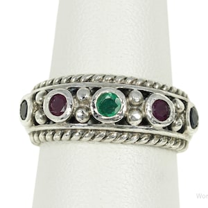 May include: A sterling silver ring featuring a central green gemstone flanked by two red gemstones and two dark blue gemstones. The band has a rope-style border on the top and bottom edges with small silver spheres between each bezel-set stone.