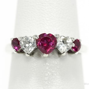 May include: A silver ring featuring five heart-shaped gemstones. Alternating between clear and deep red stones, the ring is set on a silver band. The gemstones are arranged in a row across the top of the ring.