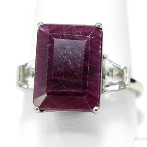 May include: A silver ring featuring a large, rectangular, deep red gemstone as the focal point. Two smaller, clear, rectangular stones flank the main stone. The ring is set against a white background, highlighting the jewelry's design.