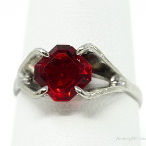 Puede incluir: Anillo plateado con una llamativa gema roja oscura de forma octogonal. El diseño del anillo incorpora garras estilizadas que sujetan la gema, creando una pieza de joyería única y llamativa.