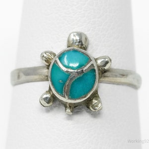May include: A silver ring featuring a turquoise turtle design. The turtle's shell is inlaid with bright turquoise stones, and the ring has a simple, polished band. The turtle's head and flippers are also silver.