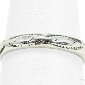 May include: A silver ring with an engraved pattern. The ring features a repeating design of oval shapes and small, triangular accents. The ring is shown on a white background.