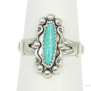 Puede incluir: Anillo de plata con una piedra turquesa. El anillo tiene una piedra central turquesa ovalada, rodeada de detalles plateados. La banda tiene un diseño decorativo. El anillo se muestra sobre una superficie blanca.