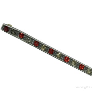 May include: A silver-coloured bar pin featuring a line of alternating red and clear gemstones. The rectangular pin has a textured border and a clasp on the back. A vintage-style jewellery accessory.